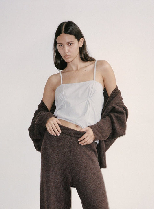 A woman stands against a plain background wearing the Pleat Top, paired with loose brown knit pants and a matching brown cardigan draped off her shoulders, looking confidently at the camera.