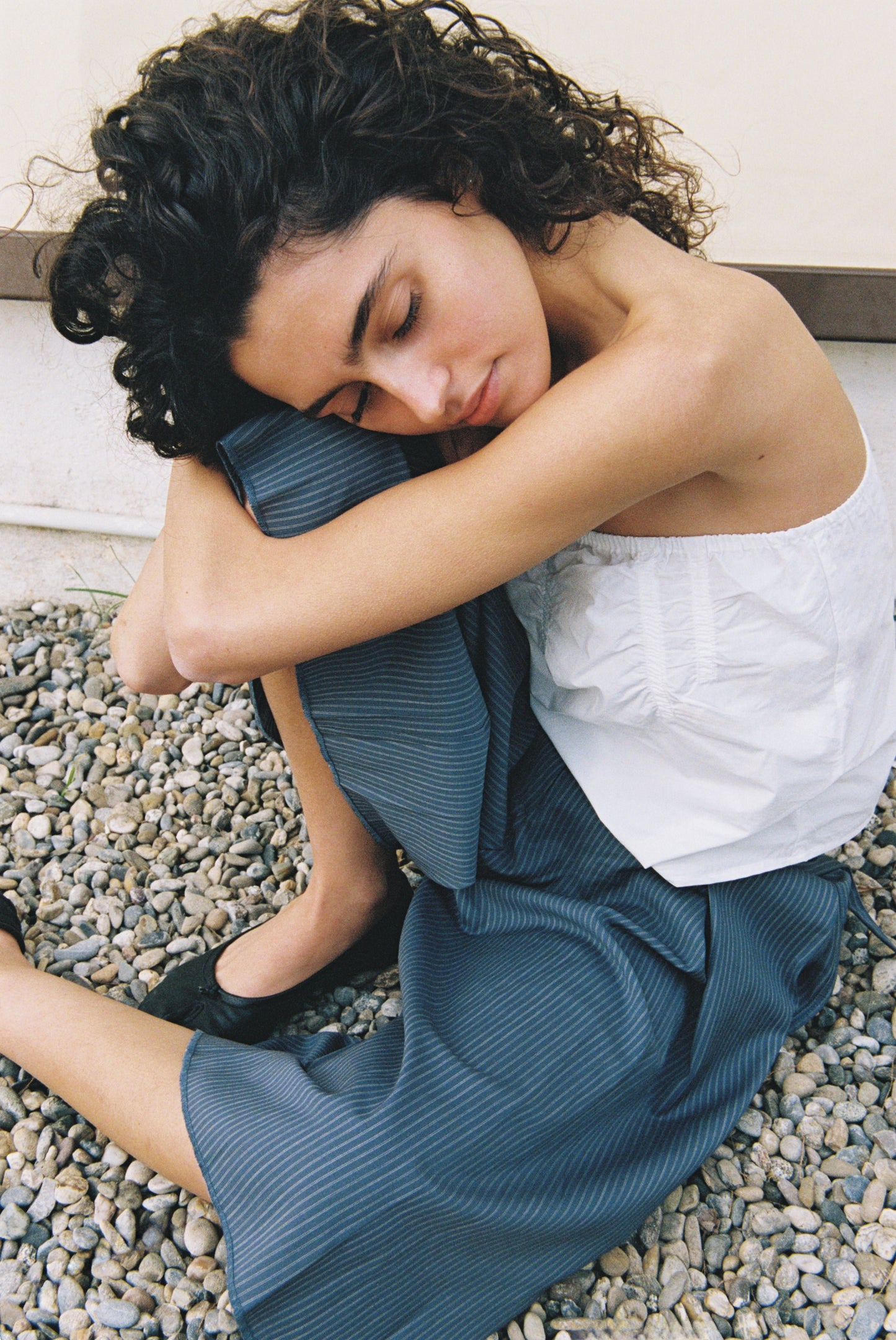 A woman with curly hair sits on gravel, hugging her knees and resting her head on them. She wears the Tube Top - White, paired with a blue skirt, appearing peaceful and relaxed with her eyes closed.