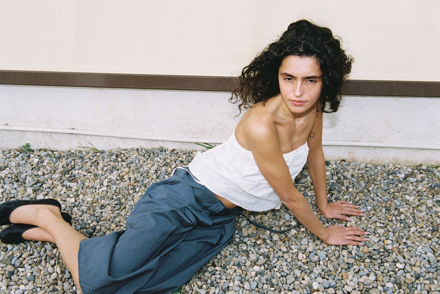 A person with long, curly dark hair wears the Tube Top - White and loose blue-gray pants while sitting on gravel, leaning back on their hands. They have black shoes, a neutral expression, and a light wall behind them.