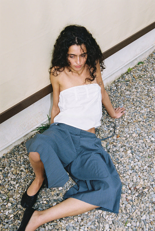 A woman with curly hair, wearing a white Tube Top and blue skirt, sits outdoors on gravel against a light wall, gazing slightly upward.