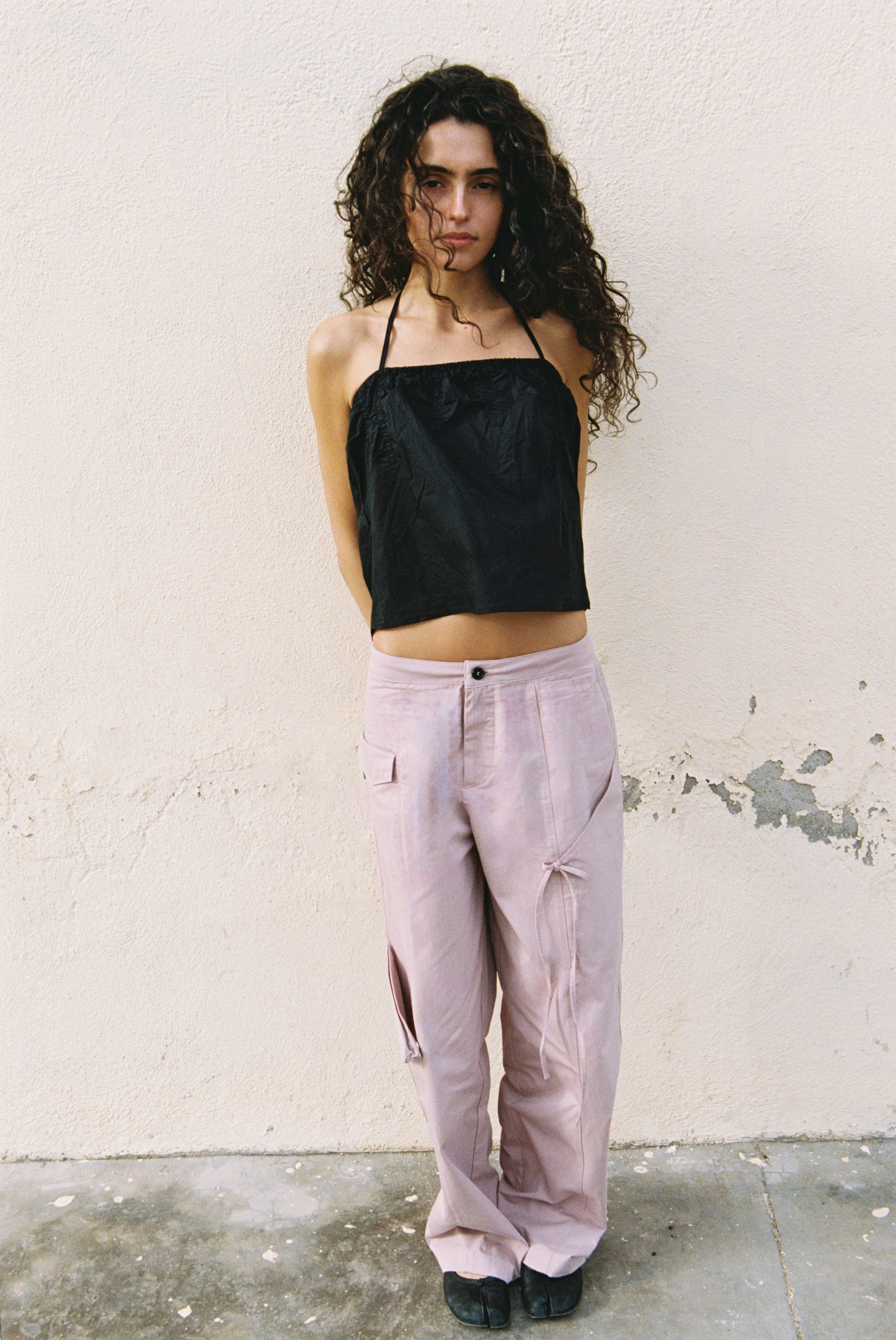 A person with long curly hair stands against a light wall, wearing a black halter top and loose pale pink Pocket Pant - Musk made from organic cotton, with their hands behind their back.