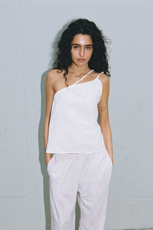 A woman with long curly hair stands against a light gray wall wearing the Shoulder Top, a white one-shoulder piece paired with loose, light striped pants. Her hands are in her pockets and she has a neutral expression.