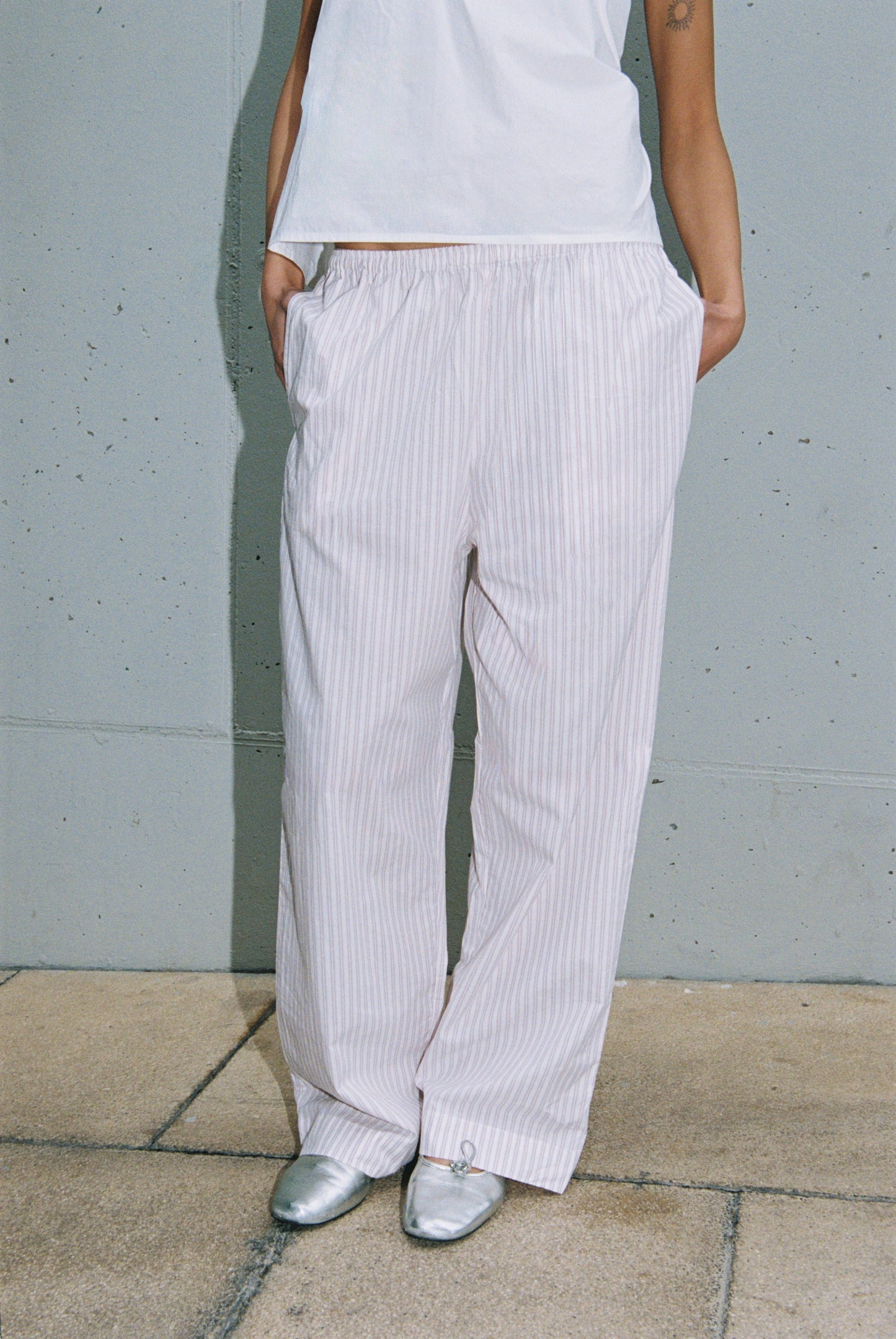 Shown are the lower legs of a person with hands in pockets, wearing Deiji Studios Ease Trouser—loose, light pink pinstriped pants—paired with a white T-shirt and silver shoes against a gray wall.
