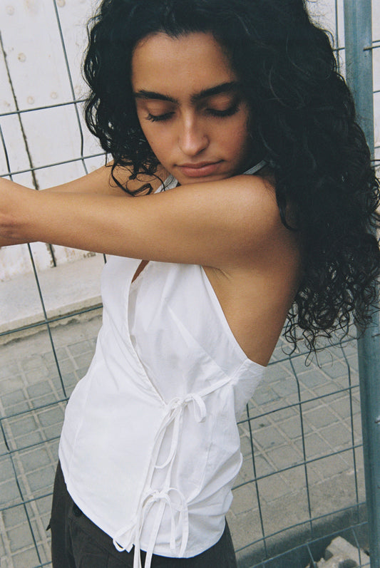 A woman with long, curly dark hair stands sideways with eyes closed, wearing the Drawcord Halter - White by Deiji Studios and dark pants, posing in front of a wire fence and tiled pavement.
