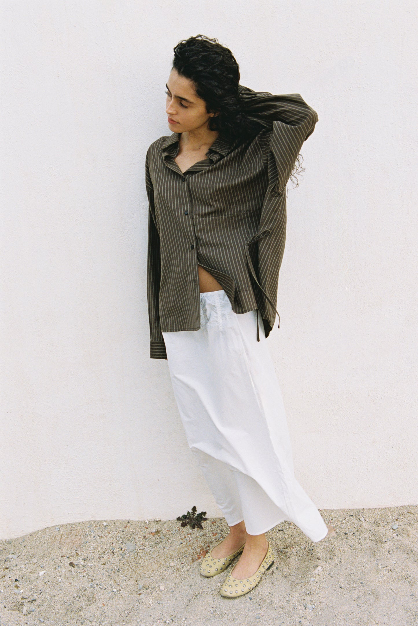A person with curly dark hair stands against a white wall, wearing the Pocket Wool Shirt—a loose brown striped top. They pair it with a long white skirt and light flats, looking down as they brush their hair back with one hand.