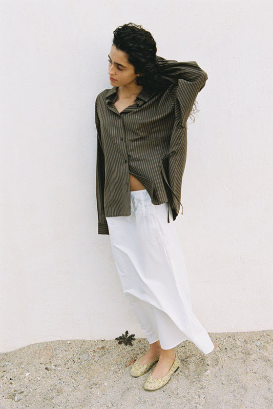 A person with curly dark hair stands against a white wall, wearing the Pocket Wool Shirt. They pair it with a long white skirt and light-colored flats, looking down while raising one hand to their hair.