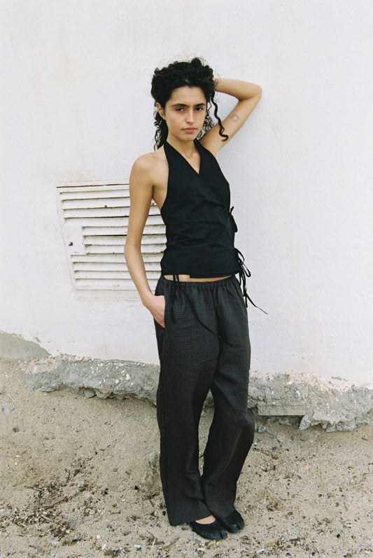 A person with curly dark hair poses against a white wall, wearing a sleeveless black top and the Linen Ease Trouser. They have one hand on their hip, a neutral expression, and stand on rough sandy ground.