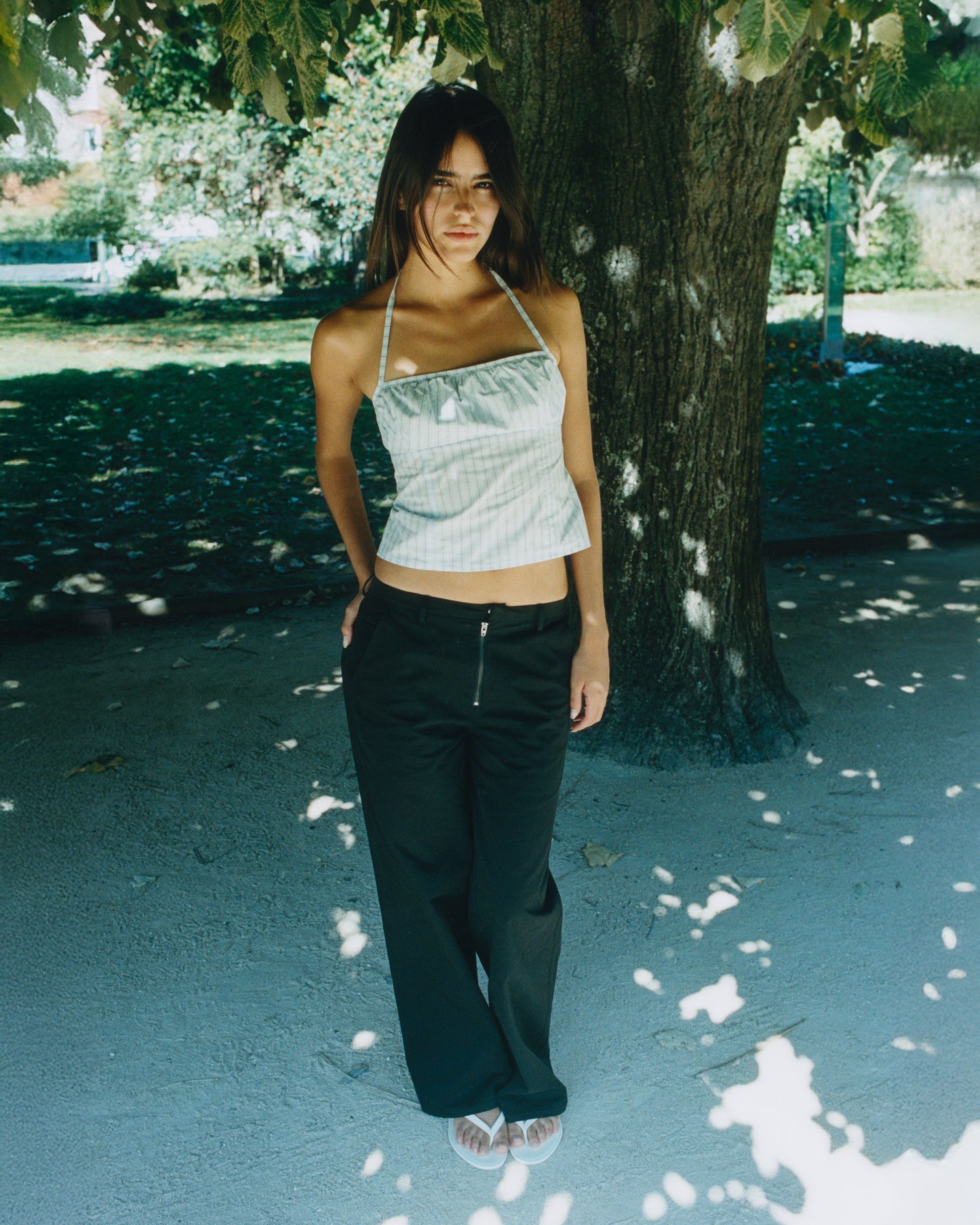 A woman stands under a leafy park tree, wearing a light-colored halter top and the Zip Pant - Black made from organic cotton twill. Dappled sunlight highlights her outfit, casting soft shadows on the ground.