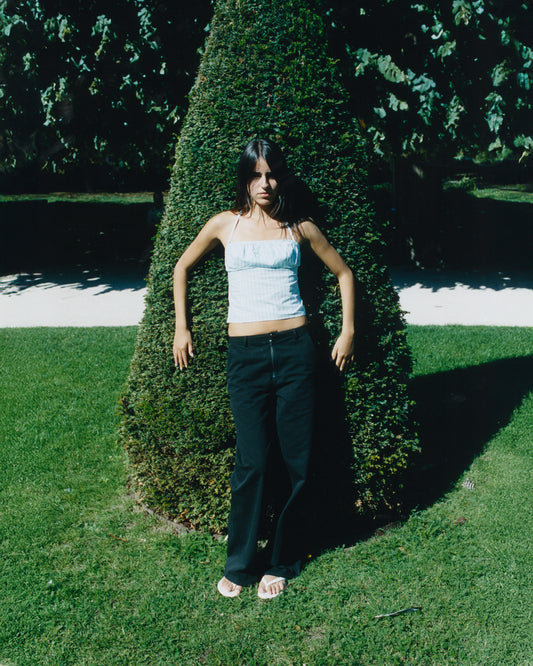 A person with long dark hair stands on a sunny lawn by a cone-shaped bush, wearing a light blue sleeveless top, Zip Pant - Black by Deiji Studios, and white sandals. Tree shadows are visible in the background.