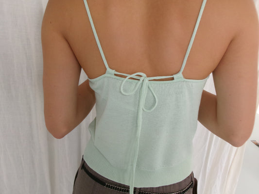 A person wears the Deiji Studios Drawcord Knit Top - Mint with thin straps and a bow tie at the back, featuring an adjustable ruched neckline. They stand against a white fabric backdrop, highlighting the open-back design.