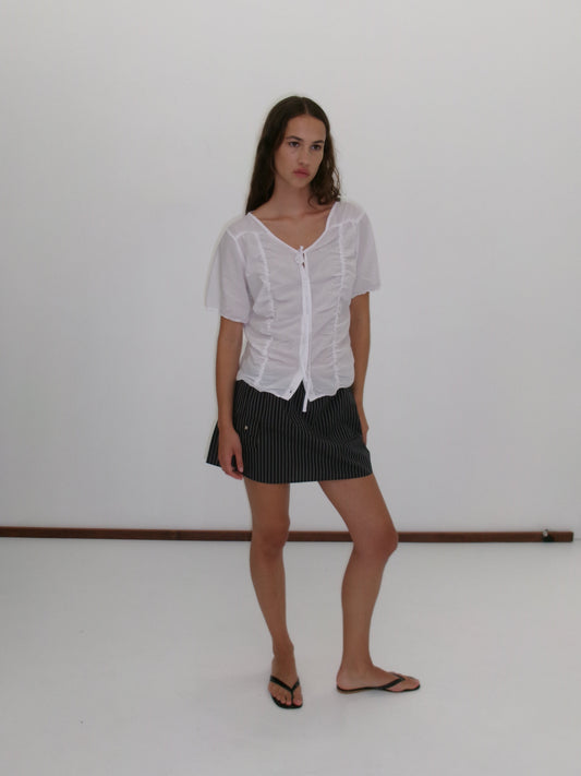 A woman with long brown hair and a neutral expression stands against a plain white background, wearing the Crinkle Top - White by Deiji Studios, a black pinstripe mini skirt, and black flip-flops.
