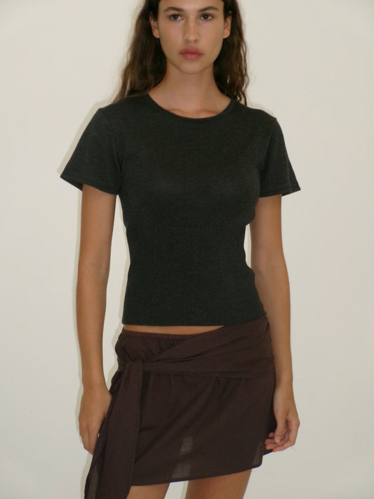 A woman with long brown hair wears a short-sleeved, fitted black top and the Belted Skirt - Wine from Deiji Studios, standing against a plain, light-colored background.