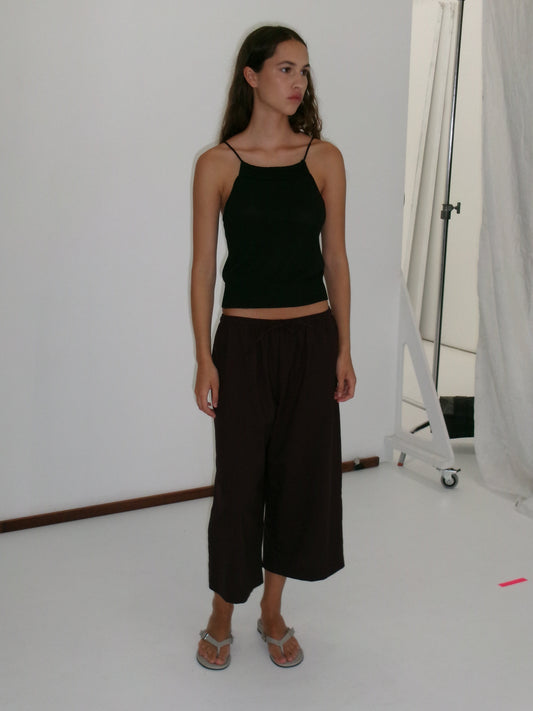 A young woman stands in a studio wearing a black sleeveless top and the Drawcord Culotte - Burgundy Brown Stripe from Deiji Studios with flip-flops. She has long hair, looks to the side neutrally, against a plain white background.