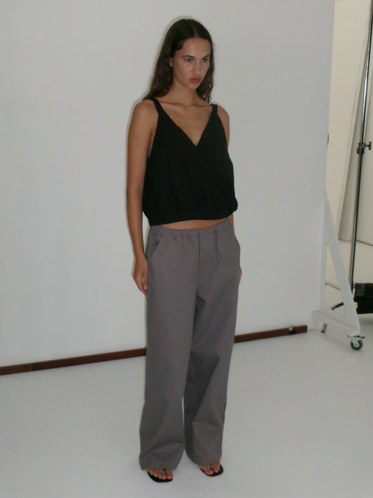 A woman with long hair and a neutral expression stands in a studio against a white background, wearing a sleeveless black V-neck top, Curve Pant - Lavender Smoke in a relaxed fit and mid-rise waist, and black sandals.