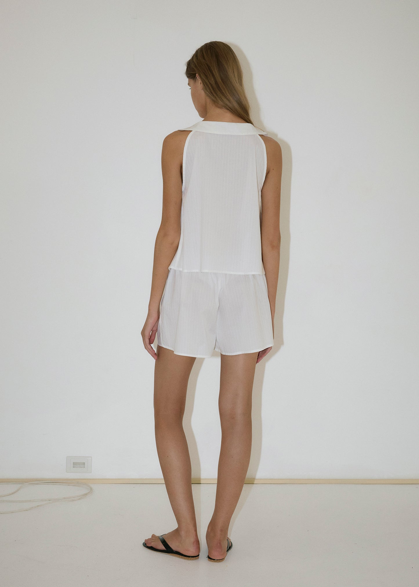 A person with long hair stands barefoot facing a white wall, wearing the Round Collar Set - White Texture. A power outlet and cable are visible near the floor.