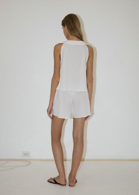 A person with long hair stands by a plain white wall, wearing the Round Collar Set - White Texture with an elastic waist and black sandals in a minimal space with a bare floor and visible outlet.