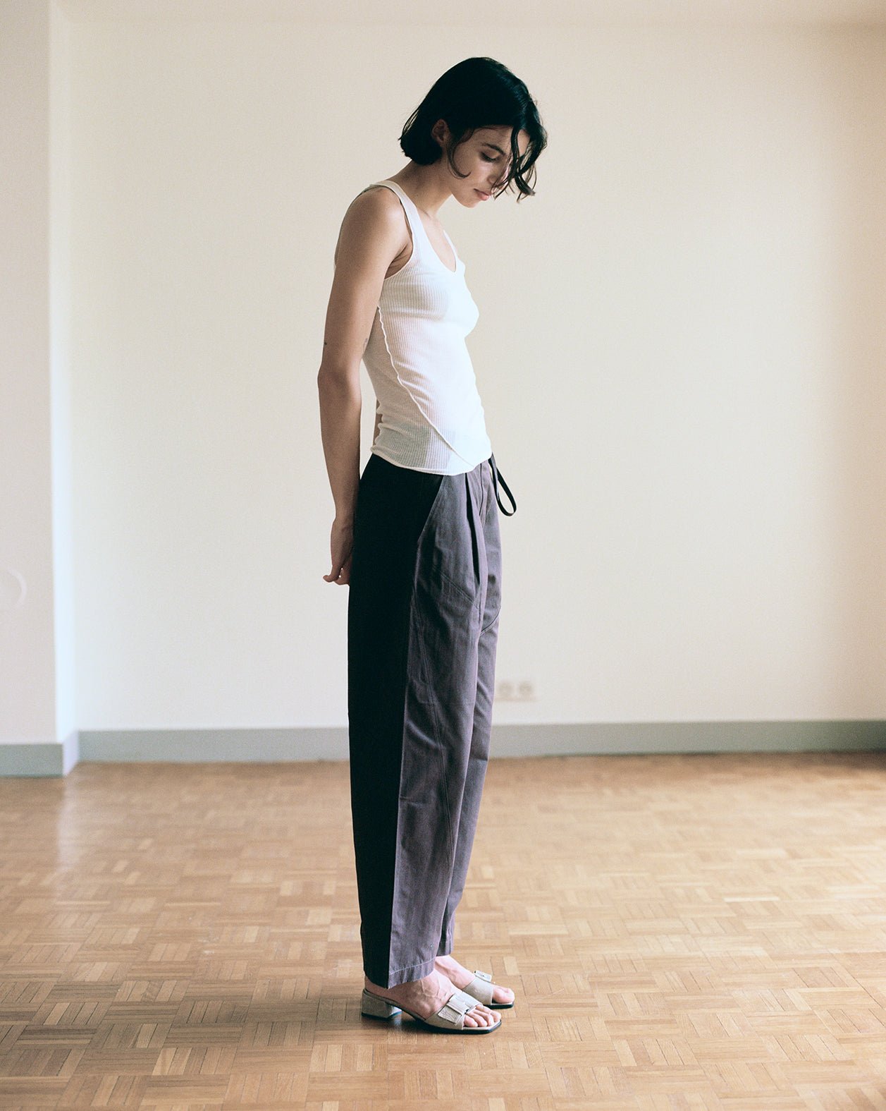 A person stands on a wooden floor in a plain room with light walls, wearing the Knit Tank - Off White, loose dark pants, and sandals, looking down with hands behind their back.