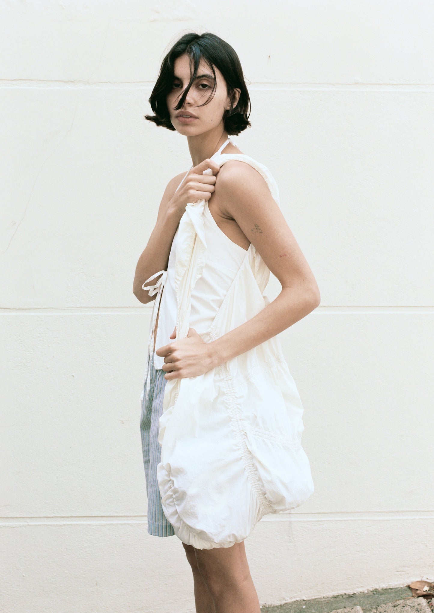 A person with short dark hair, wearing a sleeveless white ruched dress, stands against a pale wall with a neutral expression, holding the Parachute Bag—a large ruffled white bag—over their shoulder.