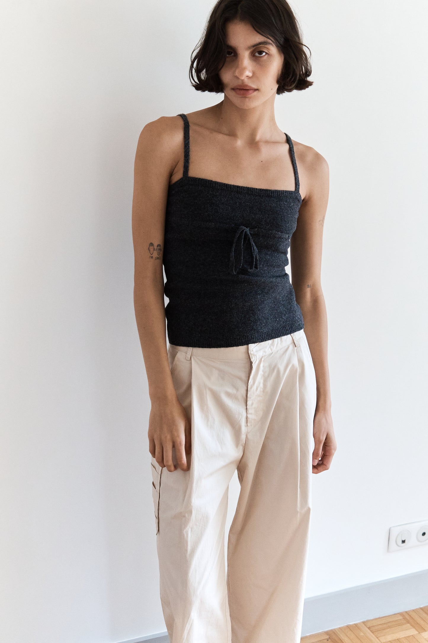 A woman with short dark hair stands against a white wall, wearing a dark gray sleeveless top and Gathered Pocket Pant in Off White organic cotton twill. She has a relaxed expression and a small tattoo on her left arm.