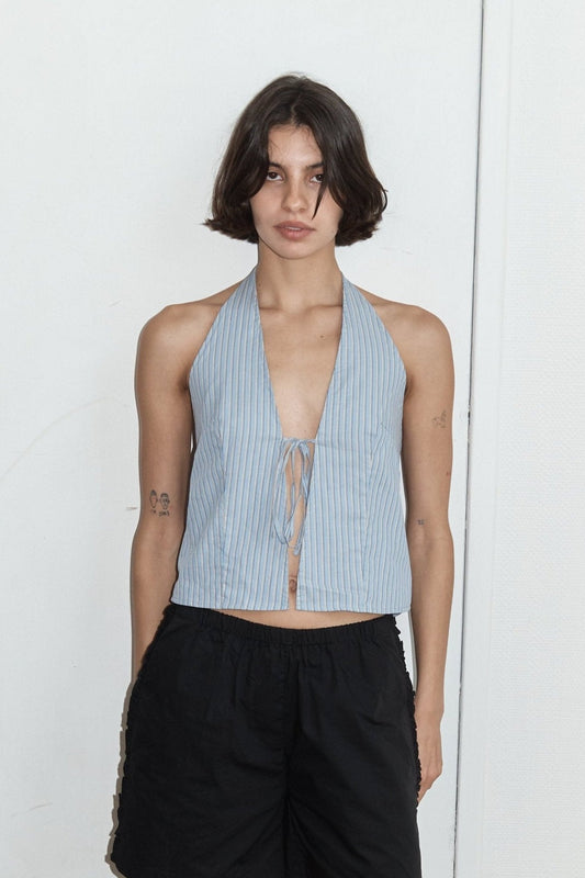A person with short dark hair models the Open Halter Top, a sleeveless light blue piece with thin vertical stripes, paired with black shorts and set against a plain, light background.
