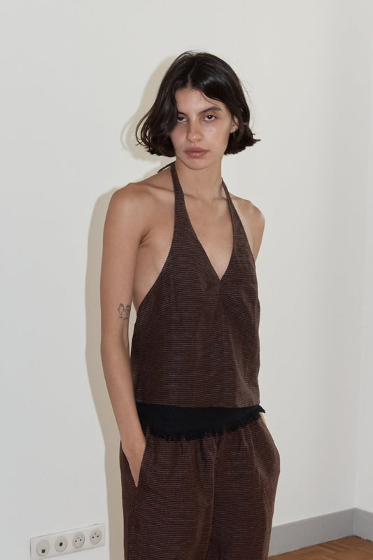 A woman with short dark hair wears a Linen Halter Top and matching pants, standing indoors against a plain white wall with one hand in her pocket.