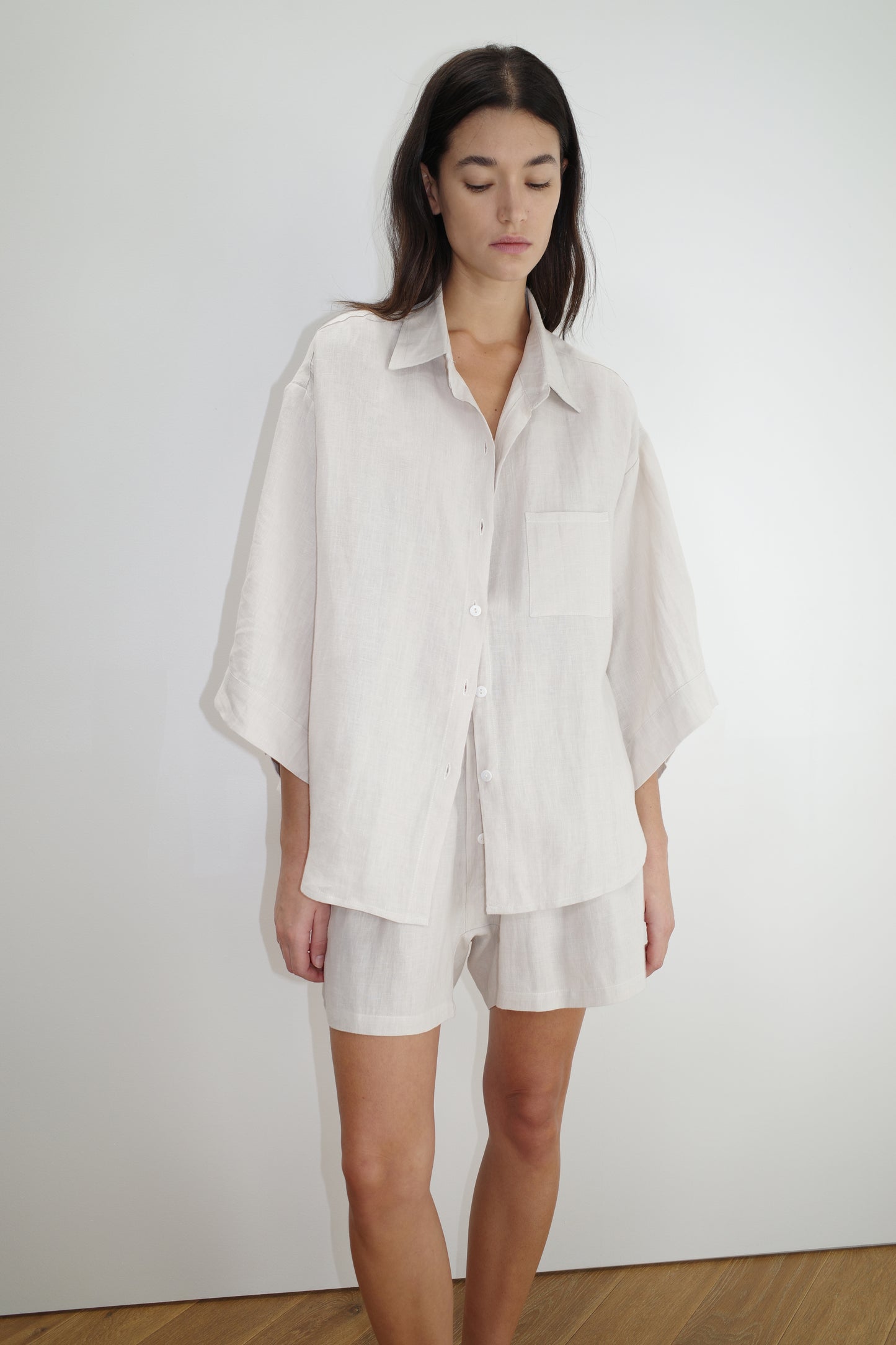 A woman with long dark hair stands against a plain white wall, wearing the 03 set - oatmeal: an oversized shirt and matching shorts made from organic French linen. She looks down with a neutral expression.