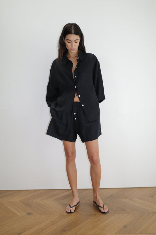 A woman stands indoors against a white wall, wearing Deiji Studios the 03 set in deep black—an oversized linen shirt with matching boxer-style shorts—paired with black flip-flops. She has long dark hair and looks down, hands in her pockets.
