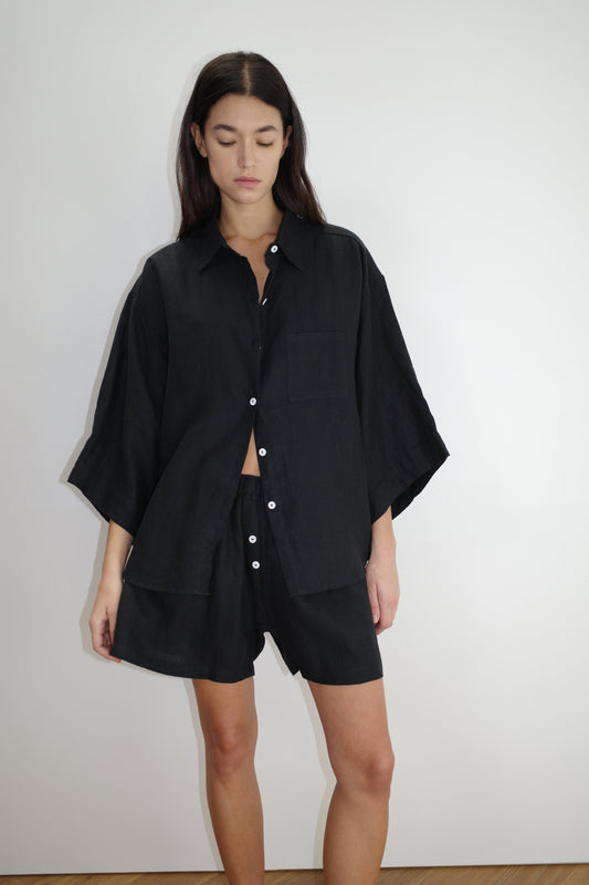 A woman with long brown hair models Deiji Studios’ the 03 set in deep black—an oversized linen shirt and matching boxer-style shorts—against a plain white background.