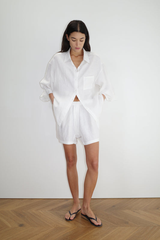 A woman stands against a white wall wearing Deiji Studios the 03 set in white—an organic linen shirt with matching boxer shorts. She has her hands in her pockets and wears black flip-flops on a wooden floor.