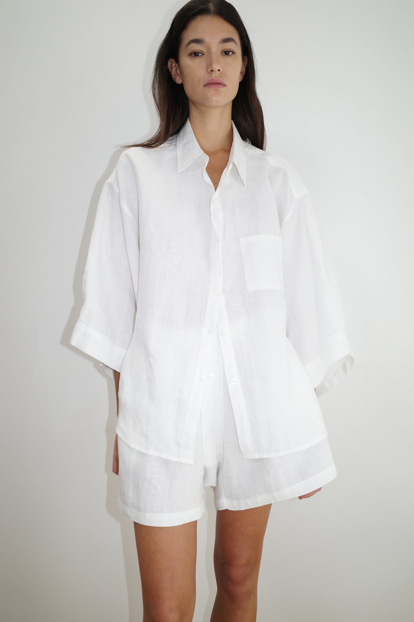 A woman with long brown hair and a neutral expression wears Deiji Studios the 03 set - white, featuring a loose-fitting organic linen shirt with a front pocket and matching boxer-style shorts, standing against a plain white background.