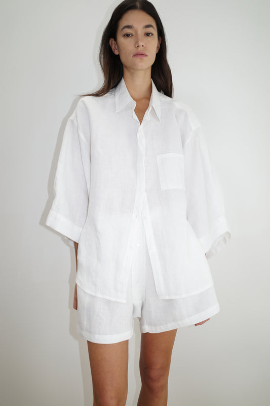 A woman with long dark hair and a neutral expression stands against a plain white background wearing the 03 set - white by Deiji Studios: a loose-fitting linen shirt and matching boxer shorts.