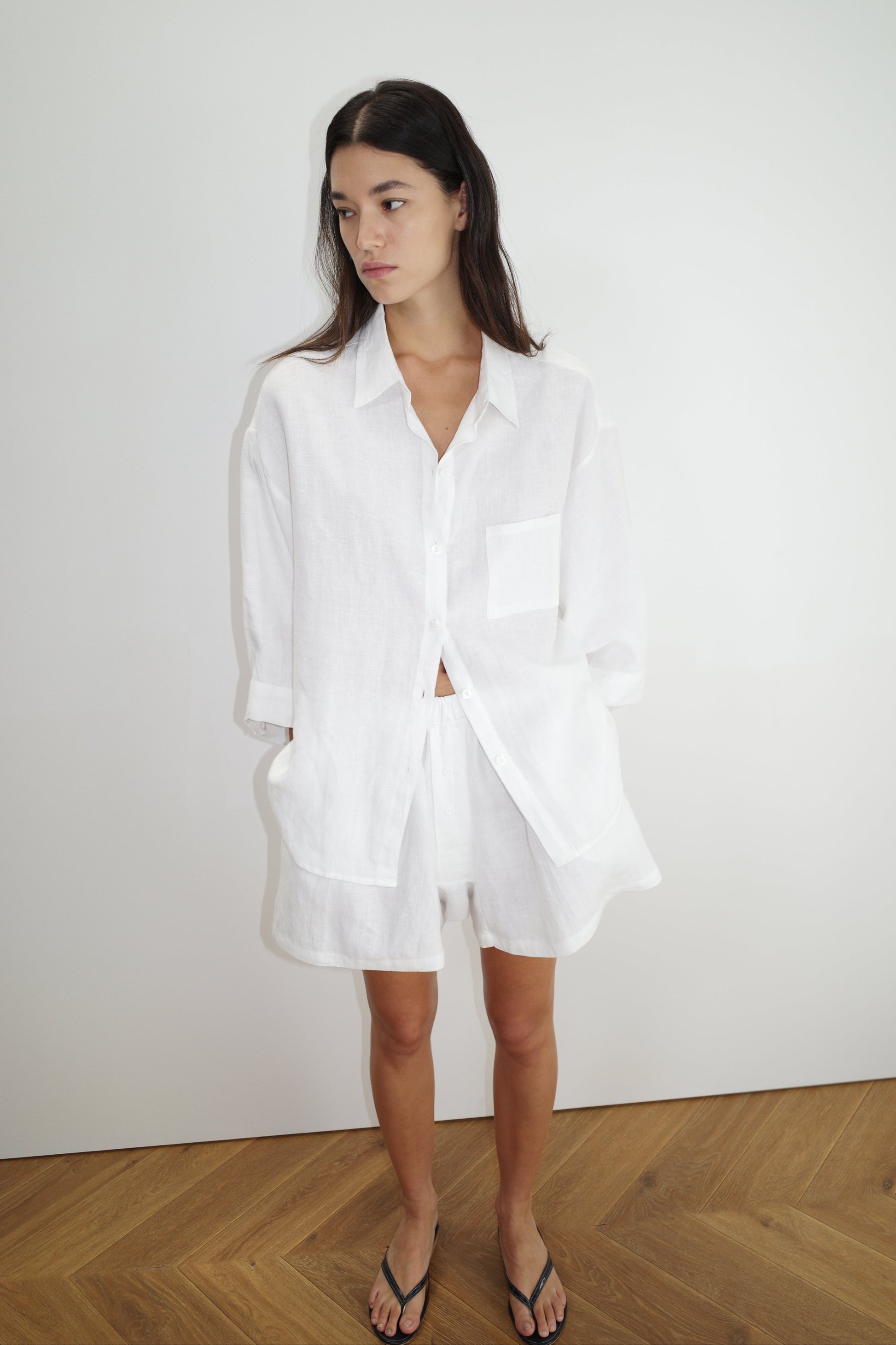 A woman with long dark hair wears Deiji Studios the 03 set in white—a loose organic linen shirt and matching boxer-style shorts with pockets—while standing indoors on a wooden floor against a plain white wall, looking to the side.
