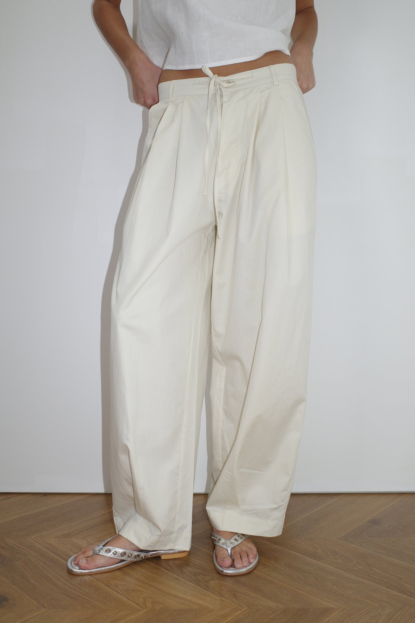 female model wears the turn pant in off white, a relaxed tailored pant features soft pleating at the front, buttoned waistband and adjustable inner drawcord. designed with hidden side seam pockets and pockets at the back, made from 100% GOTS certified organic cotton paired with a white top