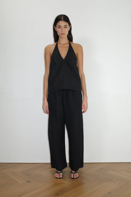 A woman stands against a plain white wall wearing loose-fitting black Linen Ease Trousers, a sleeveless black halter top, and black sandals on a wooden floor, facing forward with her arms relaxed at her sides.
