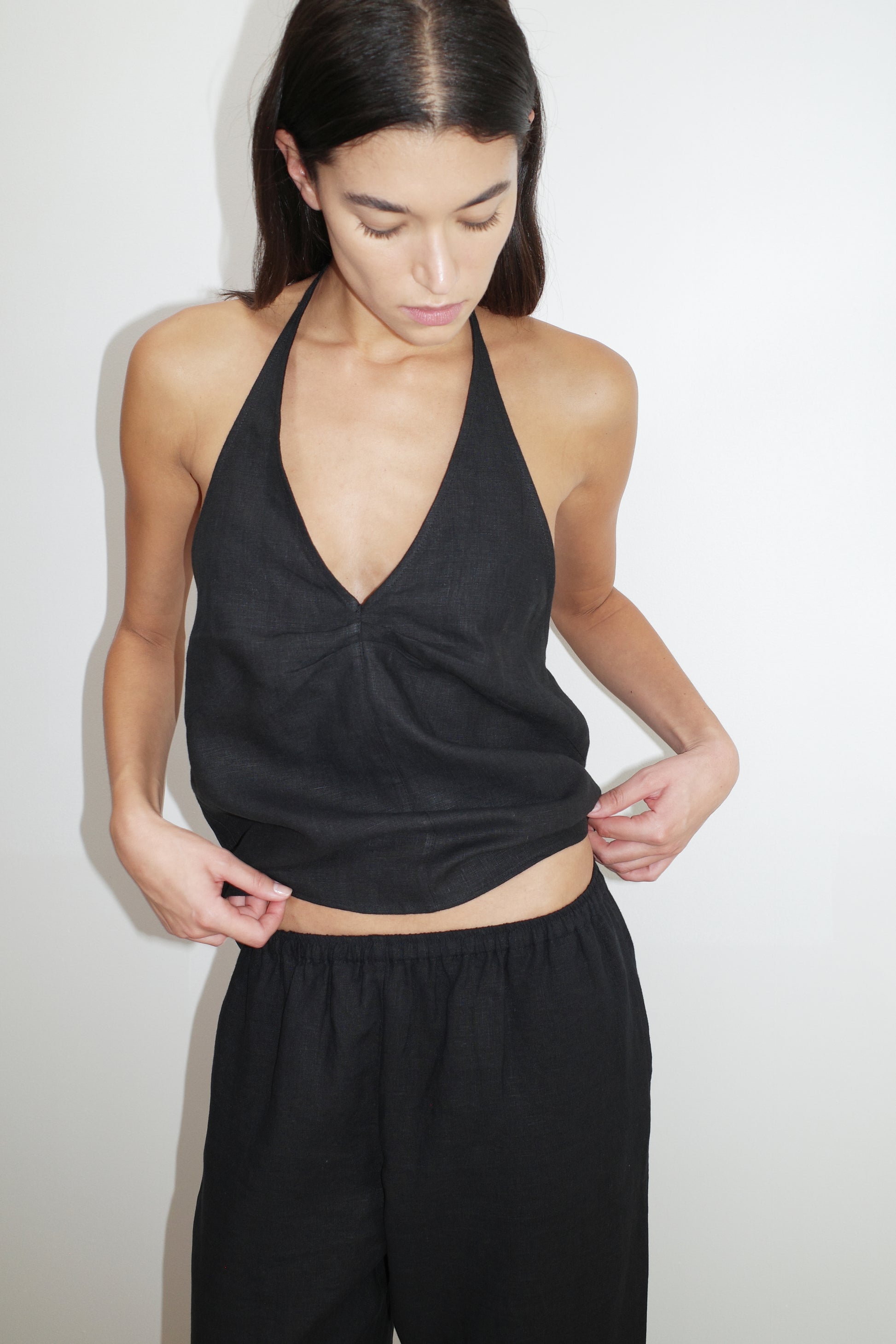 A woman with long dark hair wears the Linen Pinched Halter Top - Black and matching loose black pants, standing against a plain white background while looking down and adjusting her top.
