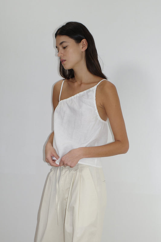 A woman with long dark hair wears the Asymmetric Tie Top - White by Deiji Studios and high-waisted cream pants, standing against a plain light background and looking slightly downward.