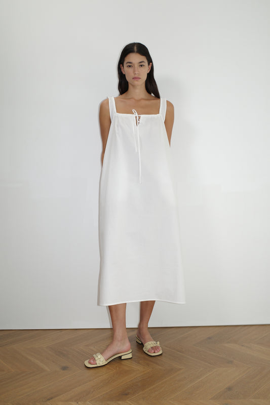 A woman stands against a white wall, wearing The Paper Dress - White by Deiji Studios with a tie neckline, paired with beige slide sandals. She stands on a light wood chevron-patterned floor.