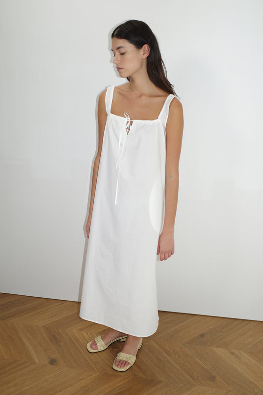 Model wearing The Paper Dress - White by Deiji Studios, featuring a square neckline, front tie, and wide straps, paired with beige woven slide sandals on herringbone wood flooring.