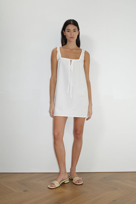 A woman stands indoors against a plain white wall, wearing The Bow Dress - White by Deiji Studios with a tie neckline and beige sandals. She has long dark hair, a neutral expression, and wooden flooring is visible.