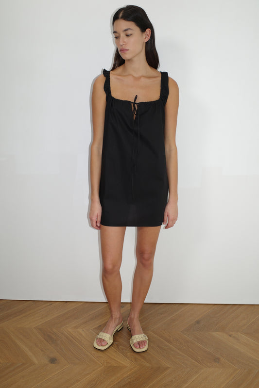 A woman with long dark hair stands indoors on a wooden floor, wearing The Bow Dress - Black by Deiji Studios, featuring ruffled straps and a tie neckline, paired with beige open-toe sandals. She is looking slightly downward.