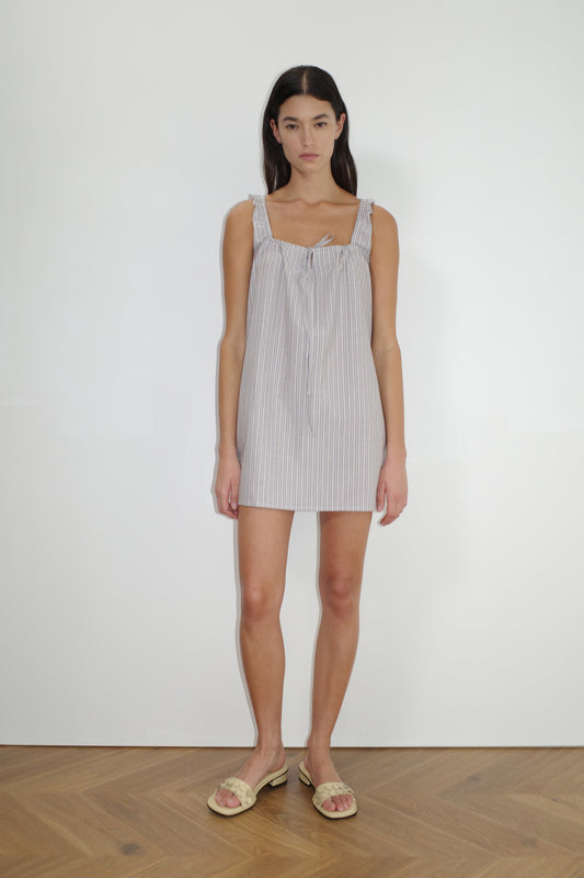 A woman with long dark hair stands against a white wall, wearing The Bow Dress - Seas Stripe, a mini-length organic cotton dress with striped pattern and tie strap details, paired with beige slide sandals on a wooden floor.