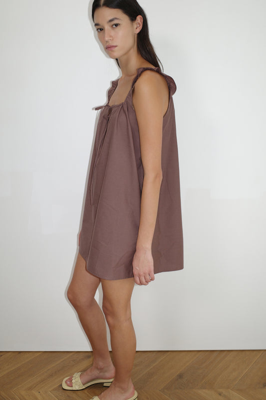 A woman stands indoors wearing The Bow Dress - Cedar, a loose, sleeveless mini dress with ruffled shoulder straps and light sandals, posed sideways against a plain white wall and wooden floor.