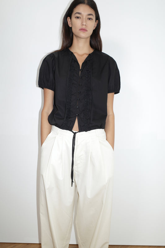 A woman with straight dark hair stands against a white wall, wearing the Double Frill Top - Black by Deiji Studios and high-waisted, loose-fitting white pants, with her hands in her pockets.