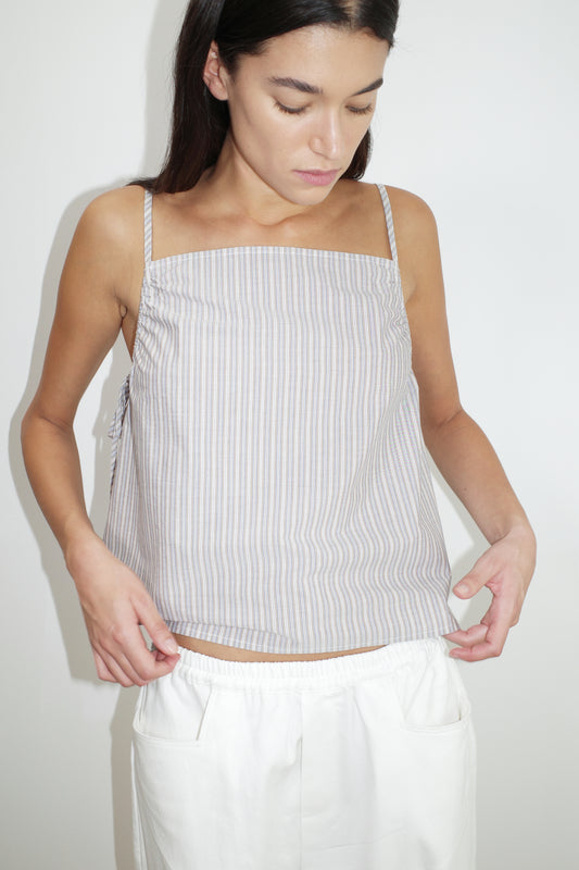 A woman with long dark hair looks down, wearing a sleeveless, square-neck Gathered Strap Top with light-colored stripes and white pants, standing against a plain white background.