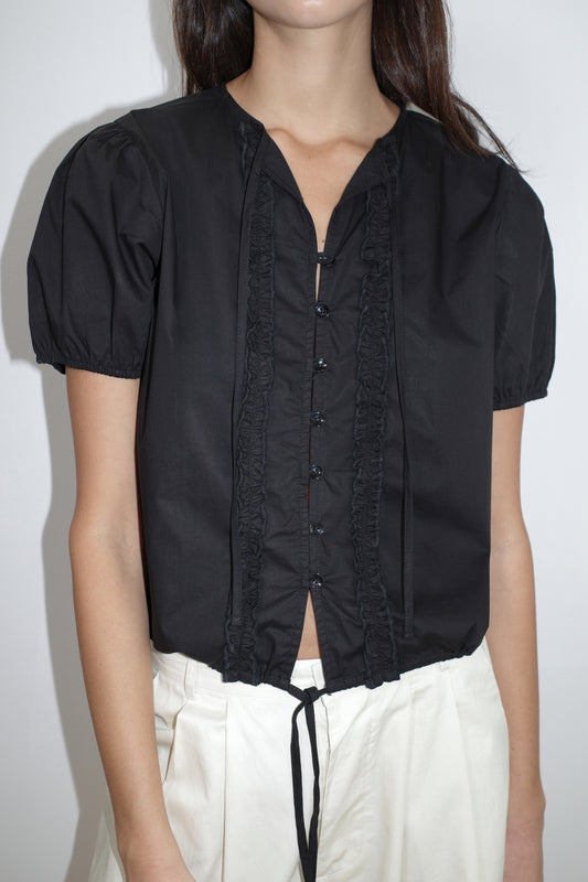 A person wears the Deiji Studios Double Frill Top - Black, a short-sleeved blouse with front buttons, paired with high-waisted loose white pants. The background is plain white.