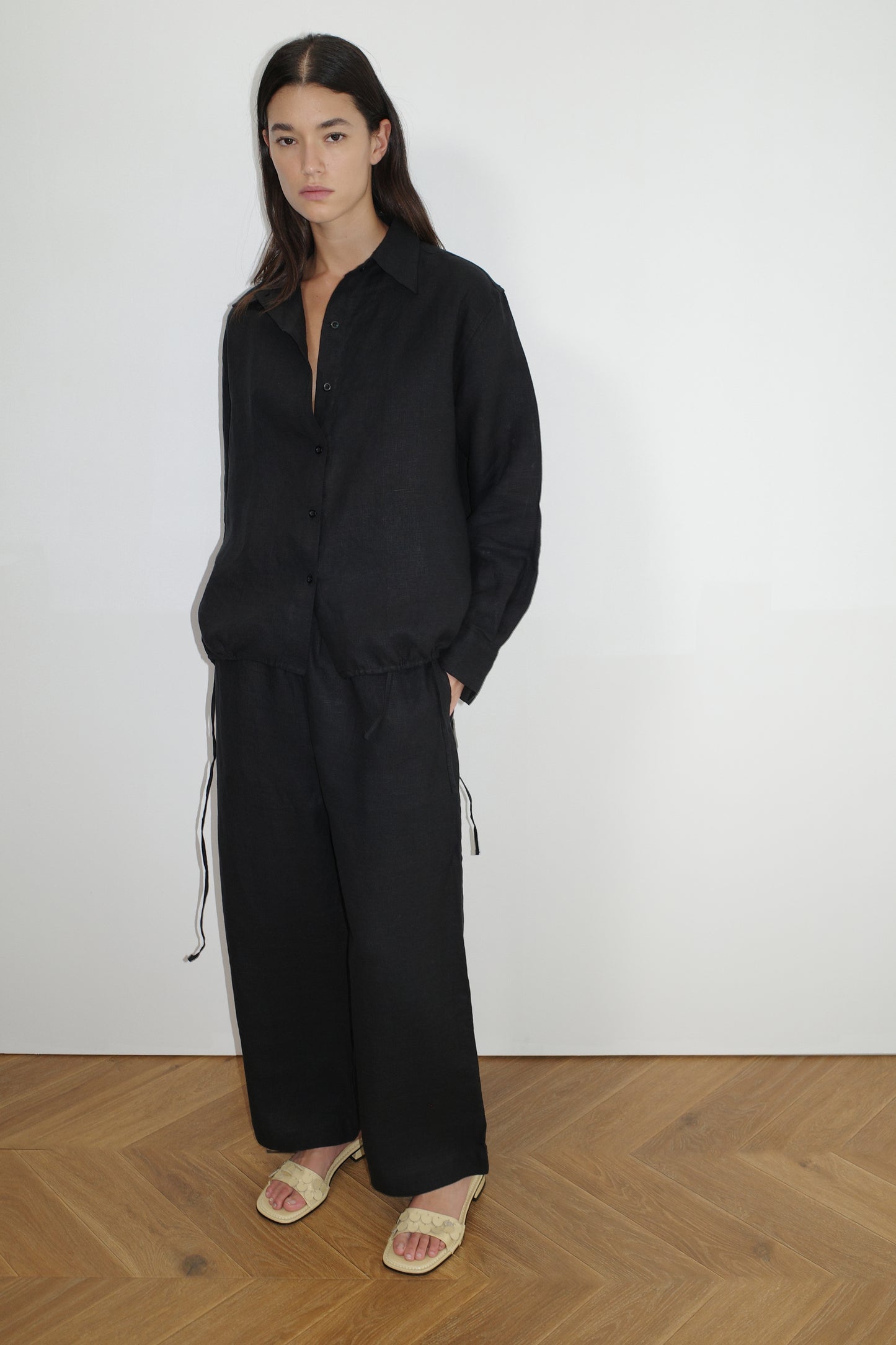 A person with long dark hair wears the Tie Split Set - Black, featuring an oversized black button-up shirt and matching wide-leg pants, hands in pockets, standing on a wooden floor against a white wall, paired with light-colored sandals.