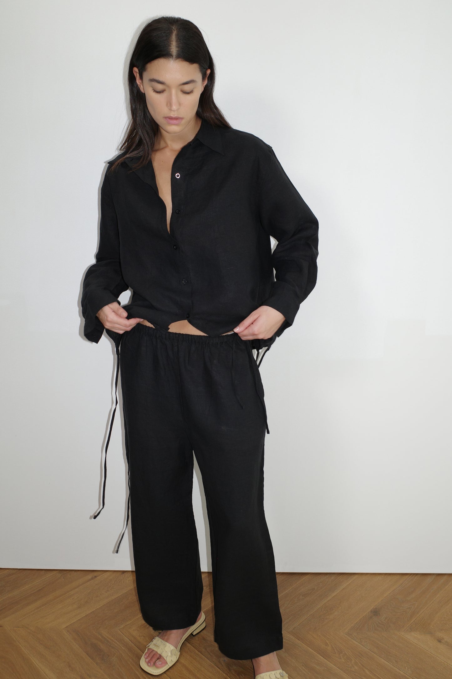 A woman with long dark hair stands indoors against a white wall, wearing the Tie Split Set - Black—an oversized black button-up shirt and matching drawstring pants—and light-colored slide sandals while looking down.