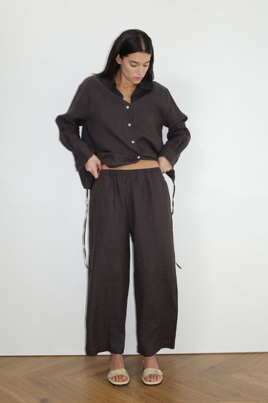 A woman stands indoors against a white wall wearing the Tie Split Set— a loose, dark brown button-up shirt with matching wide-leg pants. She looks down, hands near her waist, and completes the look with beige sandals.