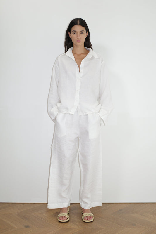 A woman stands indoors against a white wall, wearing the Tie Split Set— a loose-fitting white button-up shirt and wide-leg white pants. She has her hands in her pockets, wears light sandals, and has long dark hair.