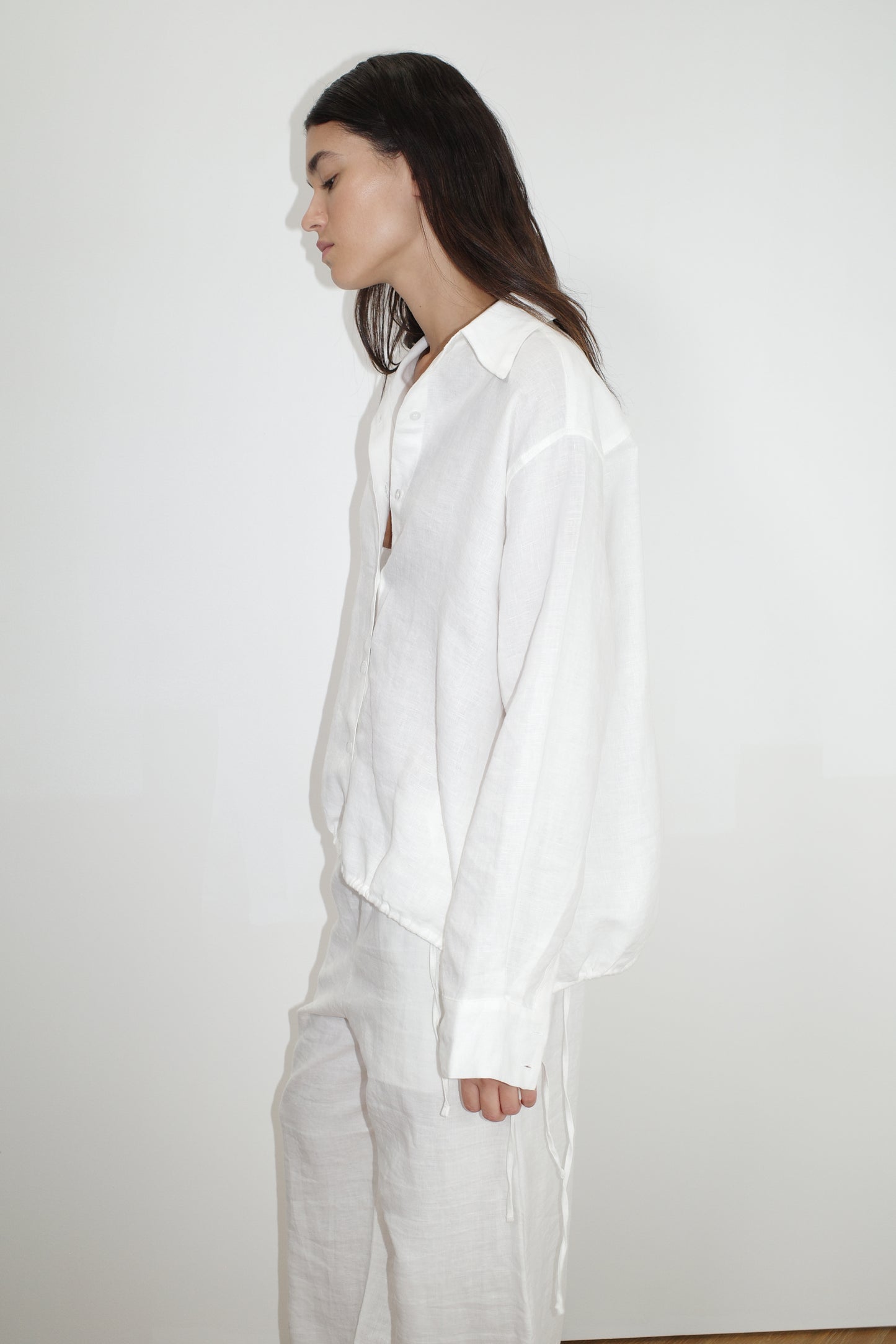 A woman with long dark hair stands in profile against a white background, wearing the Tie Split Set - White, featuring a relaxed fit organic linen button-up shirt and matching white pants.